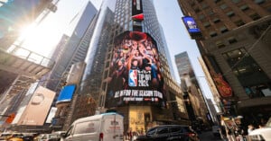 A bustling cityscape of Times Square with tall buildings and billboards. The central billboard promotes the NBA Tip-Off 2024, showing cheering fans. Vehicles and pedestrians fill the streets below, and sunlight filters through the skyscrapers.