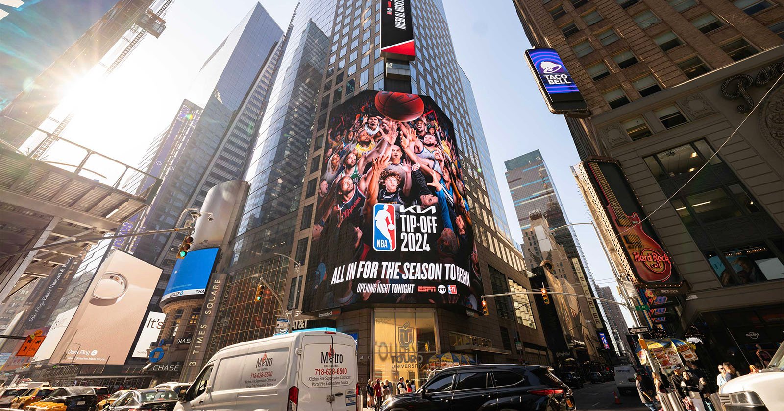 A bustling cityscape of Times Square with tall buildings and billboards. The central billboard promotes the NBA Tip-Off 2024, showing cheering fans. Vehicles and pedestrians fill the streets below, and sunlight filters through the skyscrapers.