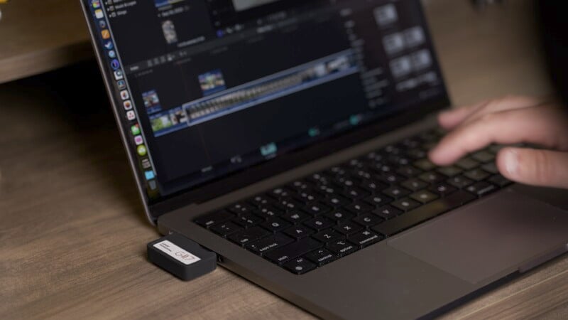 A person works on a laptop with video editing software. A small USB device is connected to the laptop, which stands on a wooden table. The person's hand is visible on the keyboard.