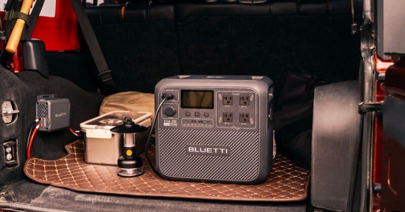 A portable power station with multiple outlets and a digital display is placed in the back of a vehicle on a carpeted mat. Nearby, there's a small light and a metal container, suggesting a camping or travel setup.