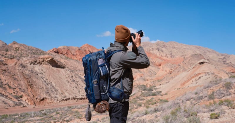 A person wearing a brown beanie and a large backpack is taking photos with a camera in a desert landscape. Rocky hills and a clear blue sky are in the background.