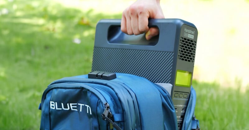 A person is placing a portable power station, labeled "BLUETTI," into a blue backpack on a grassy area. The power station features a handle and a small display screen.