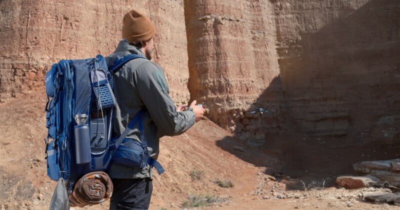 A man with a large blue backpack and brown beanie operates a remote control, flying a small drone in a rocky desert canyon. The sun casts shadows on the reddish-brown cliffs.