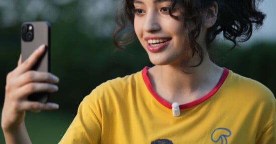 A smiling person with curly hair taking a selfie outdoors, wearing a yellow shirt with a red collar and a small pin microphone clipped to the shirt.