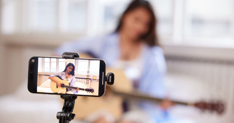 A smartphone on a tripod records a woman playing an acoustic guitar. The woman is in a softly focused background, wearing a light blue shirt, and sitting in a well-lit room.