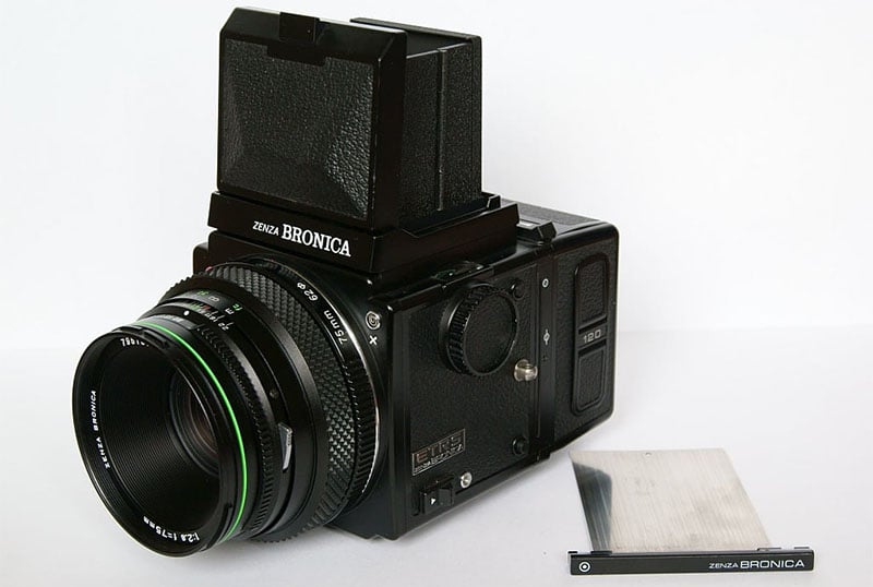 A Zenza Bronica medium format camera with a large lens and a waist-level viewfinder is displayed. The brand name is visible on the front and top. A lens cap is lying nearby on a white surface.