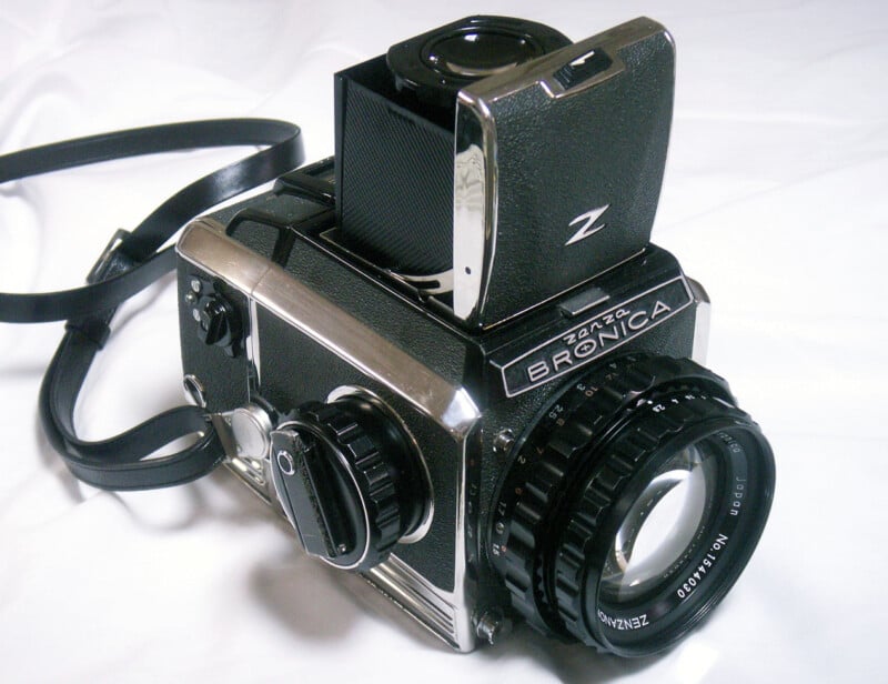 A vintage Bronica medium format camera rests on a white surface. The camera features a detachable viewfinder and a lens with visible focus and aperture rings. A black strap is attached to the camera body.