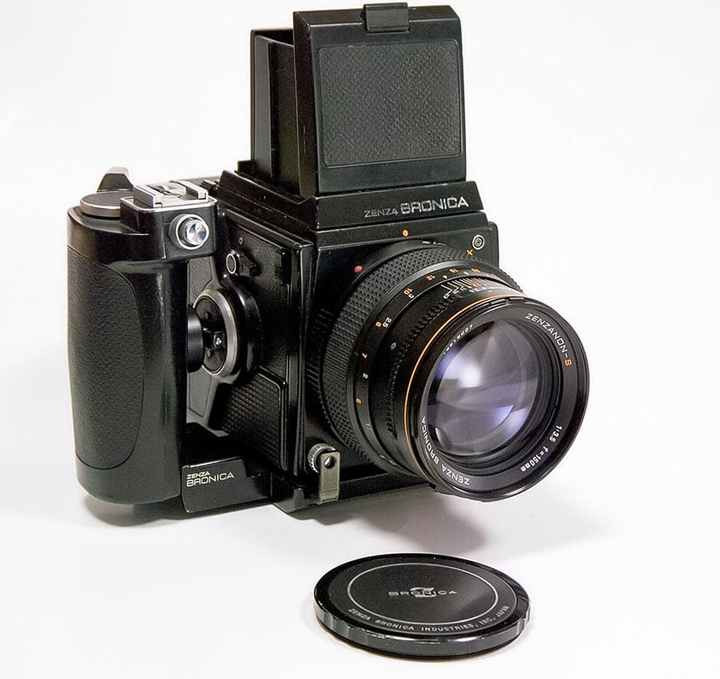 A vintage Zenza Bronica medium format camera with a large lens and a rectangular viewfinder is shown. The camera body is predominantly black. A lens cap is placed in front of the camera, resting on a white surface.