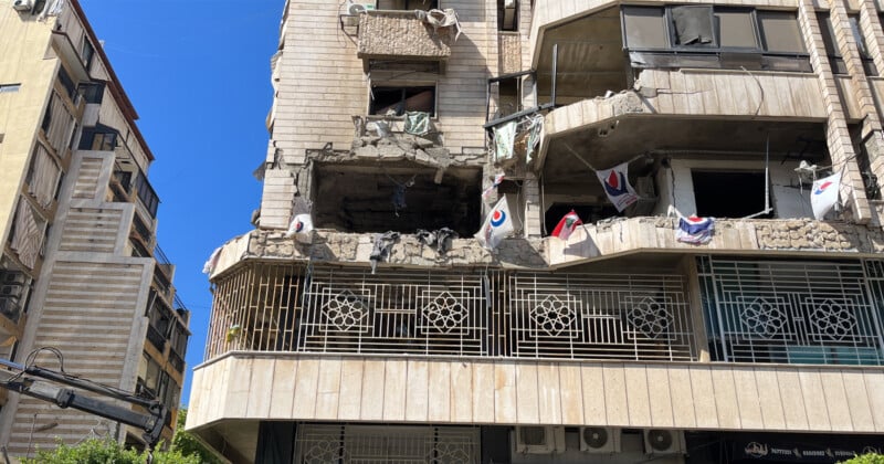 A damaged multi-story building with a partially collapsed facade. Debris is visible on the balcony, and several flags and items of clothing hang from the balcony railing. The sky is clear and blue in the background.