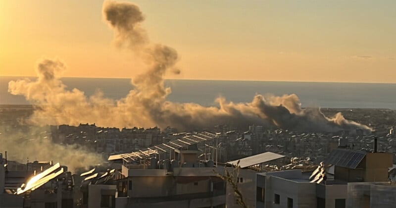 Smoke rises from buildings in a cityscape, illuminated by the setting sun. Rooftops with solar panels are visible in the foreground, and the sea stretches out on the horizon.