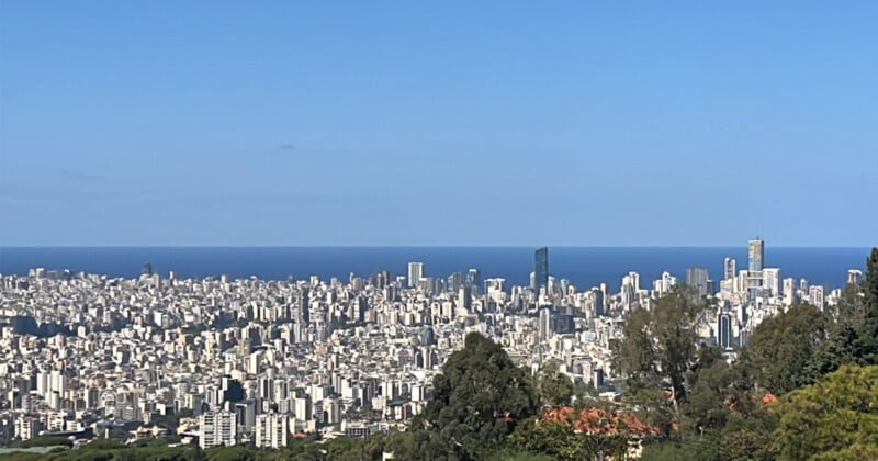 A panoramic view of a cityscape with numerous tall buildings under a clear blue sky. The city stretches towards a vast, tranquil sea in the background. Lush greenery is visible in the foreground.