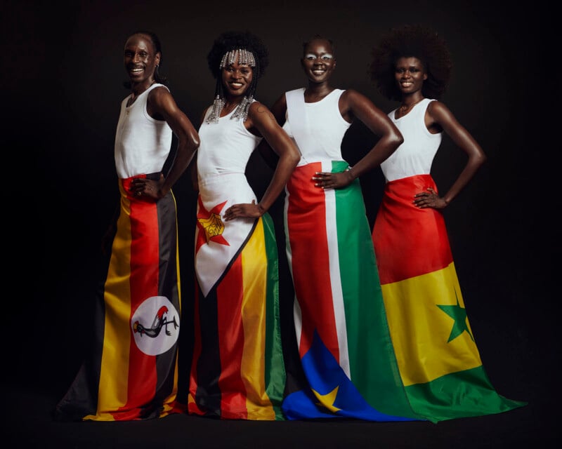 Four people stand confidently in white tops and long skirts featuring the flags of Uganda, Zimbabwe, South Sudan, and Senegal. They are smiling, posed against a dark background.