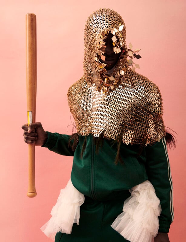 A person in a green tracksuit and white tulle skirt holds a wooden bat. Their head and shoulders are covered with a metallic gold chainmail piece adorned with small square decorations, set against a pink background.