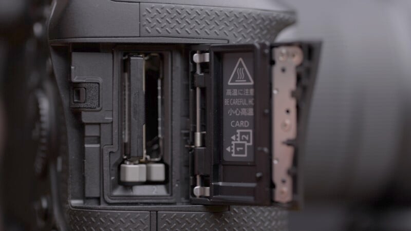 Close-up of an open camera slot showing two memory card slots. The card compartment cover is open, revealing a caution symbol and instructions. The camera body has a textured grip surface.