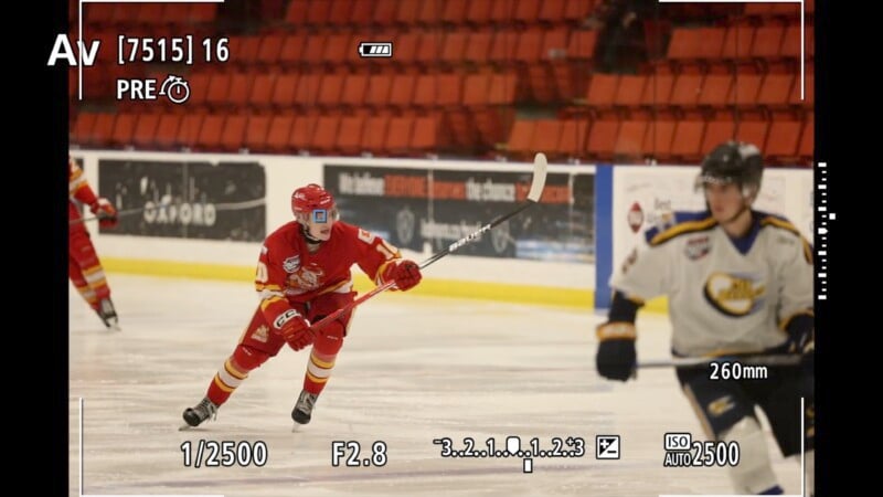 A hockey player in a red uniform skates across the rink in mid-action, holding a stick. Another player in a white uniform is nearby. The scene is framed by a camera’s viewfinder display, showing settings like ISO and shutter speed.