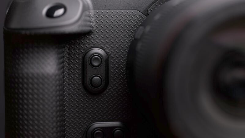 Close-up of a textured camera grip showing two circular buttons on the side of the camera body. The lens is partially visible on the right, blurred in the foreground.