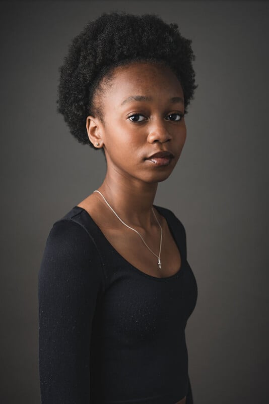 A person with short curly hair is wearing a black top and a silver necklace, standing against a neutral gray background. They have a calm expression and are looking slightly to the side.