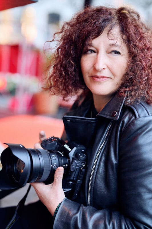 A person with curly hair, wearing a leather jacket, holds a camera while sitting outdoors. The background is softly blurred with hints of red, suggesting a casual, creative setting.