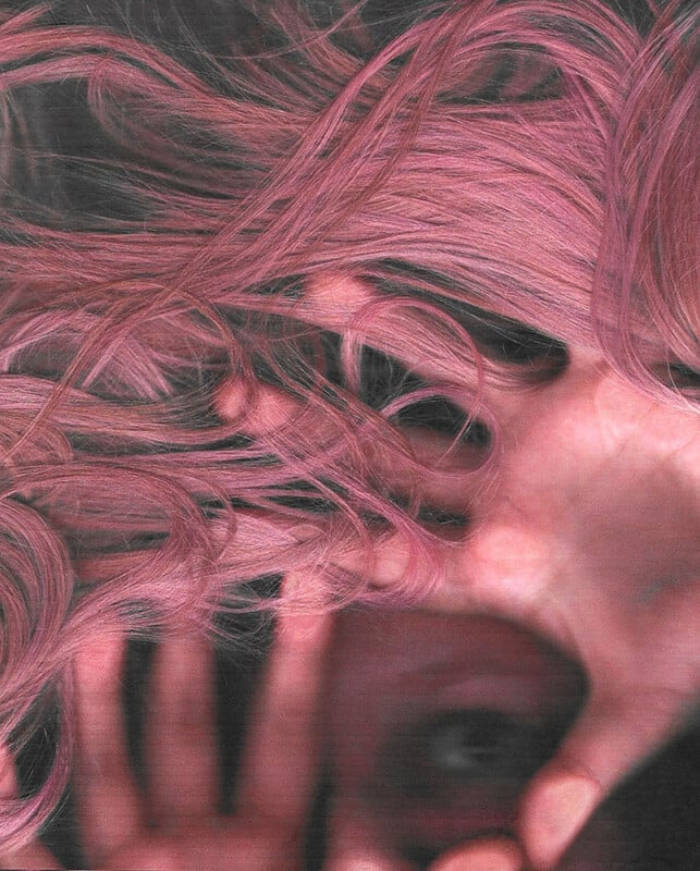 A person partially obscured by wavy, pink hair, with their hand covering part of their face. The image has a close-up and abstract feel, with a focus on the texture of the hair and the hand.