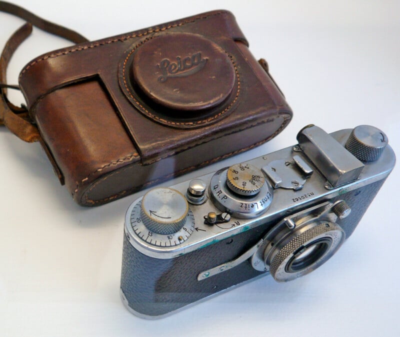 A vintage Leica camera with a textured body and intricate dials is displayed next to a brown leather carrying case. The case has a strap and embossed logo, suggesting it complements the camera's elegant design.