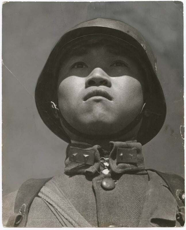 A black and white photograph of a soldier gazing upward. The soldier wears a helmet and military uniform with visible insignia on the collar. The perspective is from a low angle, emphasizing the sky in the background.