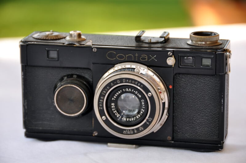 An antique Contax camera with a Zeiss lens, featuring a vintage design in black leather and metal details, is displayed against a blurred background.