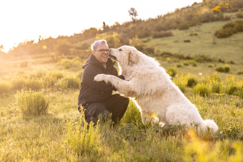 A person kneels in a grassy field, smiling and interacting with a large, fluffy white dog. The sun sets in the background, casting a warm glow over the landscape.