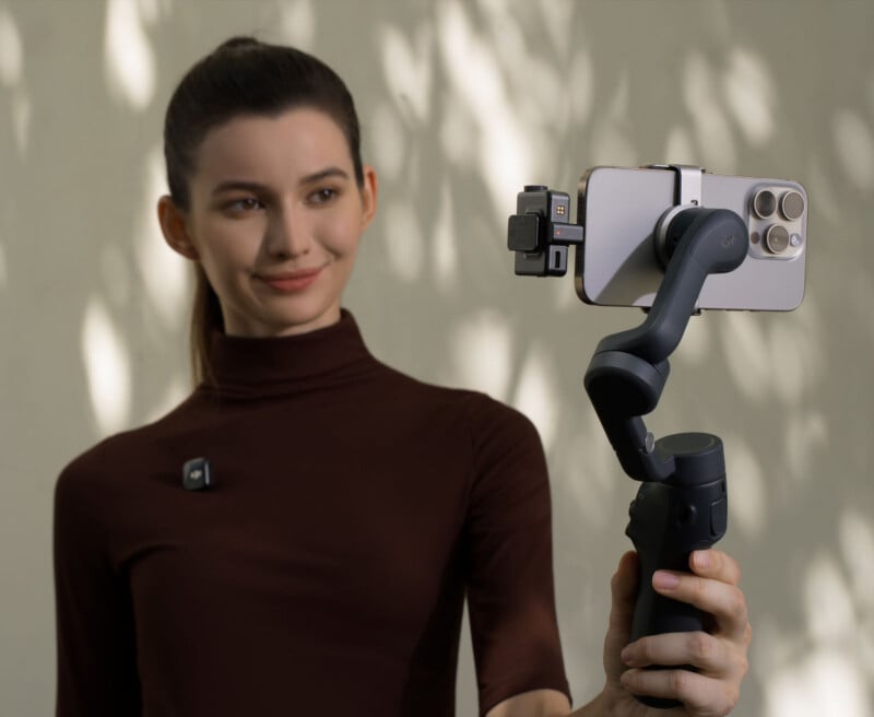 A person holding a smartphone mounted on a handheld stabilizer, appearing to be recording or taking a selfie. The background features dappled light patterns on a wall. The person is wearing a dark turtleneck and a small microphone.