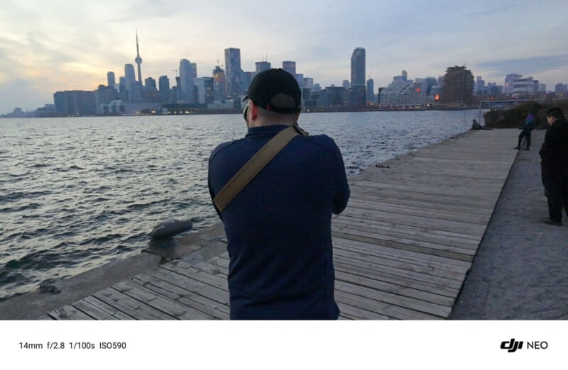 A person stands on a wooden boardwalk by a body of water, taking a photo of a city skyline at dusk. The skyline includes a recognizable tower and various buildings. The sky is cloudy with a hint of orange from the sunset.