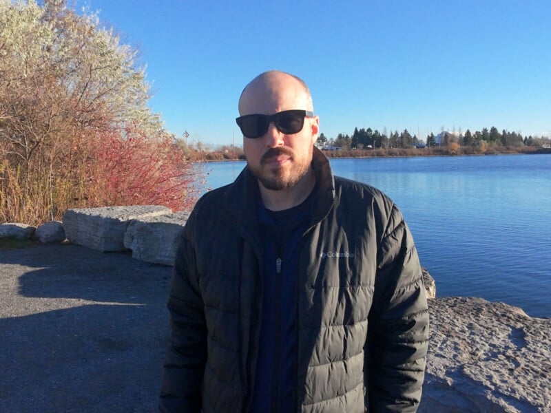 A person wearing sunglasses and a black jacket stands near a lake with a clear blue sky. There are bare trees and shrubs nearby, and rocks line the water's edge. The person has a neutral expression, and it's a sunny day.