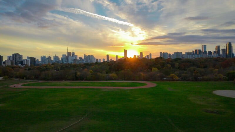 Aerial view of an expansive park with a circular path surrounded by green grass. In the background, a city skyline is silhouetted against a vibrant sunset sky with streaks of clouds.