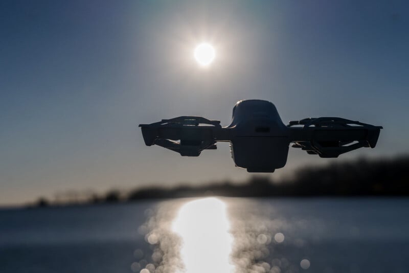 A drone hovers over a body of water during sunset. The sun is low in the sky, casting a bright reflection on the water's surface, creating a dramatic silhouette of the drone and surrounding landscape.