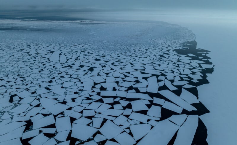 Aerial view of a vast expanse of broken sea ice floating on dark water. The ice is fractured into irregular geometric patterns, stretching toward the horizon under a cloudy sky, creating a striking contrast between the white ice and deep blue water.