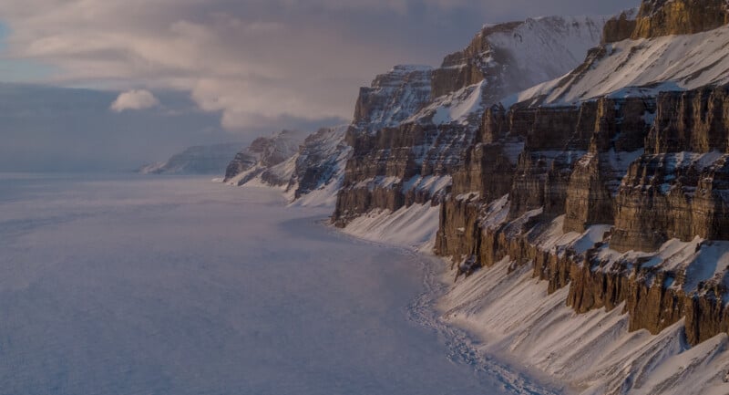 A dramatic view of a snow-covered, rugged coastline with steep cliffs bordering a vast, frozen expanse. The sky is partially cloudy, casting a serene light over the tranquil winter landscape.