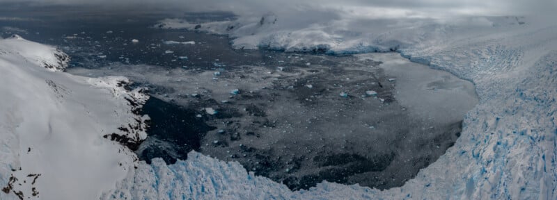 A panoramic view of a snowy and icy landscape, featuring a vast expanse of ice and snow with scattered icebergs floating on the water. Cloudy skies loom overhead, casting a gray tone over the scene.