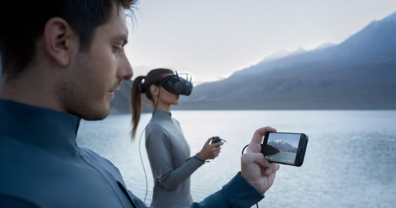 A man holds a smartphone displaying a mountain scene by a tranquil lake. In the background, a woman wearing a VR headset stands facing the water, holding a controller, with mountains visible in the distance.