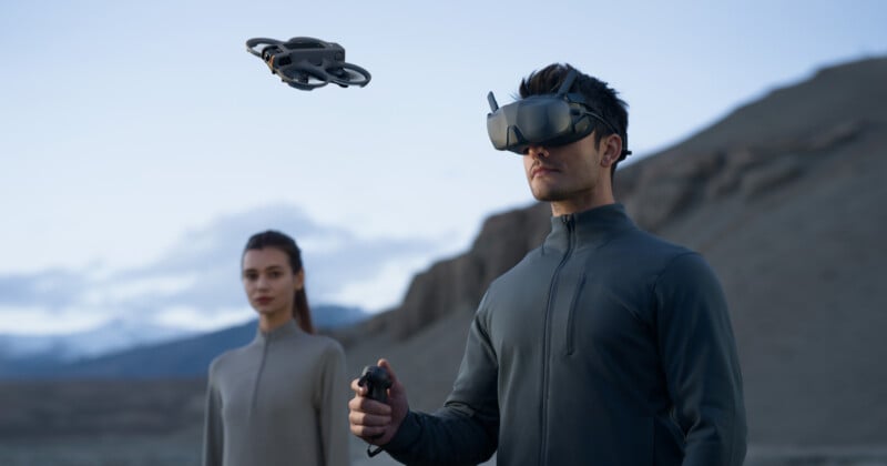 A man wearing virtual reality goggles operates a handheld controller, piloting a small drone in a mountainous landscape. A woman stands nearby, observing the drone. Both are dressed in dark, sleek clothing.