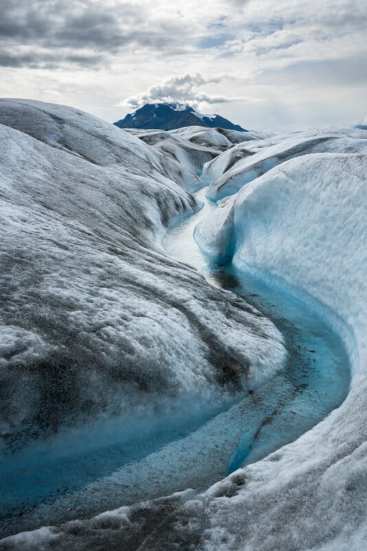 A winding stream of vibrant blue water cuts through a vast landscape of white and grey glacial ice, with a cloud-covered mountain peak in the background under a partly cloudy sky.