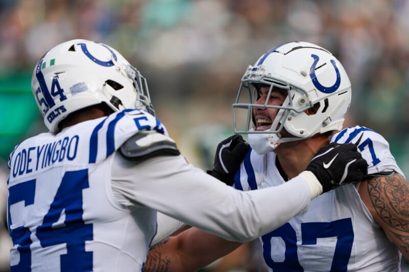 Two football players in white Indianapolis Colts uniforms celebrate on the field. One sports jersey number 54 and the other 97. Both wear helmets, and the scene captures a moment of excitement and camaraderie during a game.