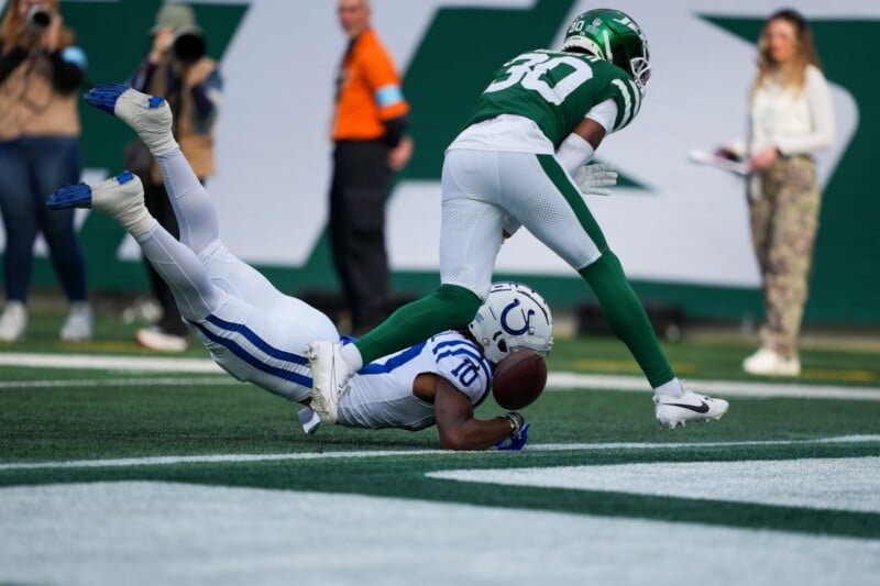 A football player in a white and blue uniform falls to the ground while holding the ball near the end zone. A player in a green and white uniform tries to block him. The scene takes place on a football field with spectators in the background.