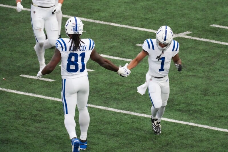 Two football players in white and blue uniforms are on a green field. One, wearing jersey number 81, reaches out to shake hands with the other, who wears jersey number 1. Both are wearing helmets, and they appear to be celebrating or encouraging each other.