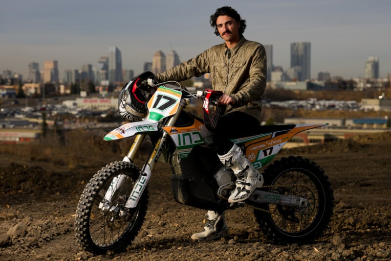 A person with a mustache is sitting on a dirt bike wearing a brown jacket and pants, holding a helmet. The city skyline is visible in the background. The bike is white and green with the number 17.