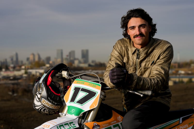 A person with a mustache sits on a dirt bike labeled with the number 17. They are wearing a brown jacket and black gloves, with a city skyline visible in the background. A helmet rests on the bike's handlebars.