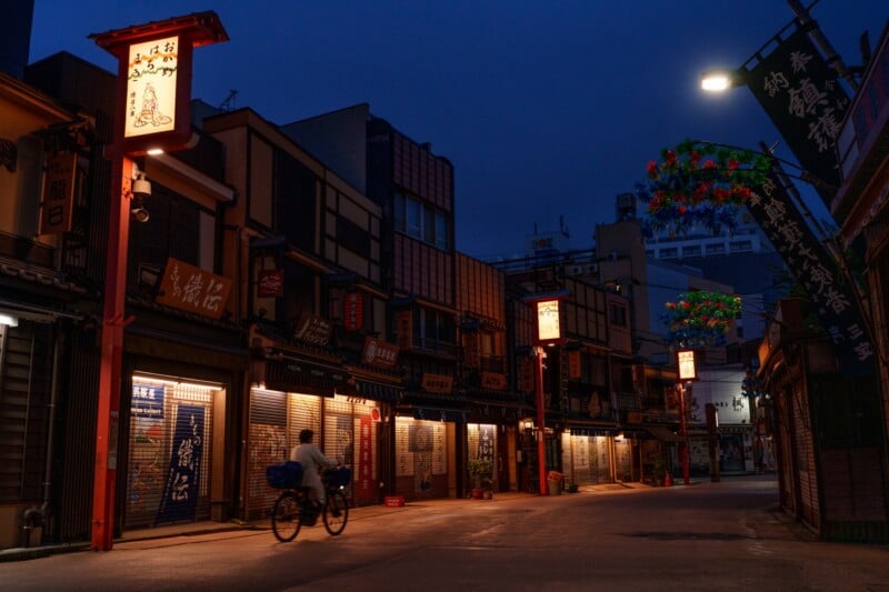 A person rides a bicycle down a narrow street lined with traditional-style Japanese shops, illuminated by warm lanterns at dusk. The sky is dark blue, and decorative lights hang overhead.