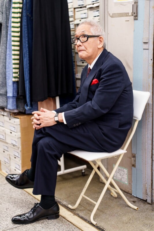 An elderly man in a navy suit and glasses sits thoughtfully on a white chair in a room filled with clothing and shoe boxes. He has a red pocket square in his jacket and shiny black shoes.