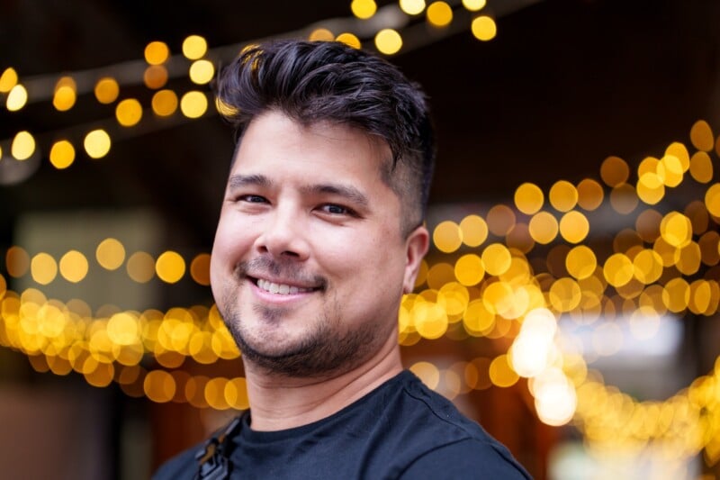 A man with short dark hair and a beard smiles at the camera. He is wearing a black shirt. The background is decorated with blurred warm yellow string lights, creating a festive and cozy atmosphere.