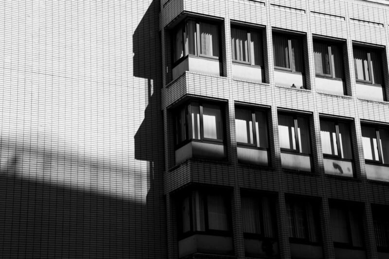 A black and white photo of a building with a grid-like pattern on the facade. It features rectangular windows, some open, casting shadows. The sunlight creates contrasting shadows on the textured surface.