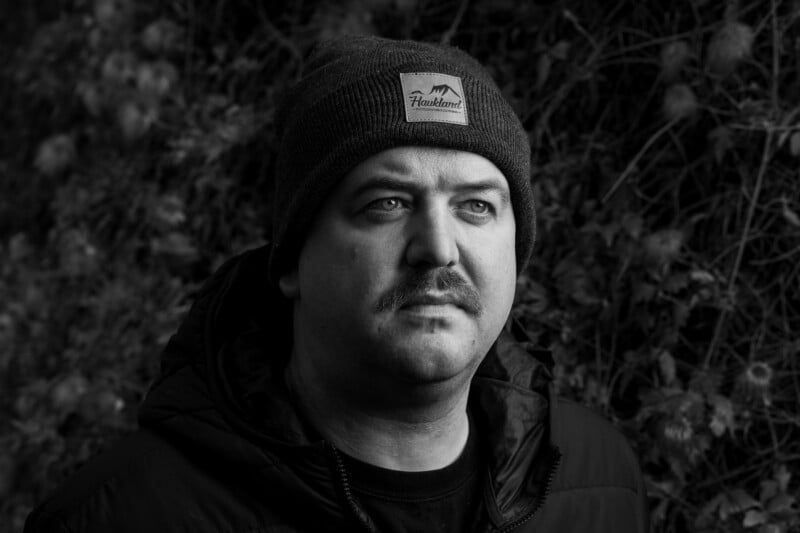 Black and white portrait of a man wearing a dark jacket and a beanie with a mountain logo. He looks slightly upward with a thoughtful expression, standing against a blurred background of foliage.