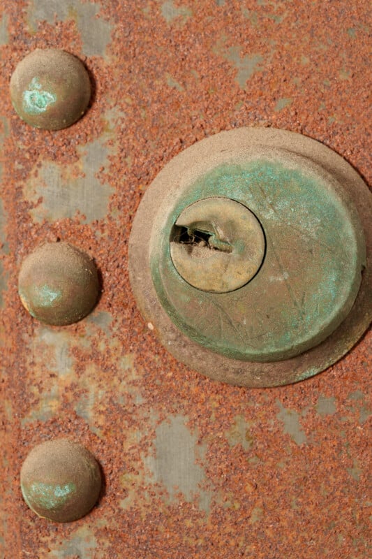 Close-up of a weathered, rusted metal surface featuring a corroded circular lock with visible green patina and three round, rusty rivets to the left.