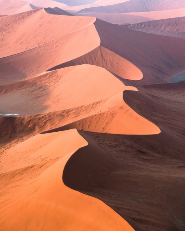 Aerial view of vast orange sand dunes in a desert landscape, creating a wavy pattern with contrasting shadows. The scene highlights the smooth curves and texture of the dunes under soft sunlight.